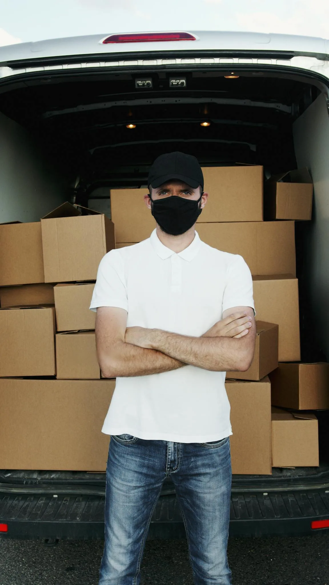 A courier wearing a mask standing in front of a loaded van with cardboard boxes, showcasing logistics.